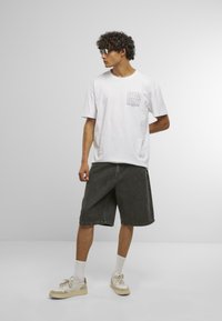 White cotton T-shirt with "SPEED DEMON" print, paired with gray denim shorts and cream sneakers. Model stands against a light background.