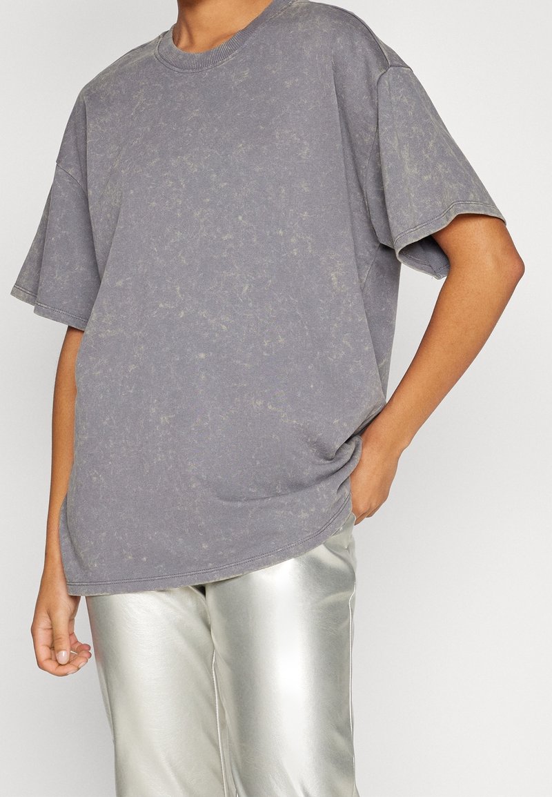 Šedé oversized tričko vyrobené z jemného materiálu, s jemným texturovaným vzorem a krátkými rukávy, doplněné lesklými stříbrnými kalhotami.