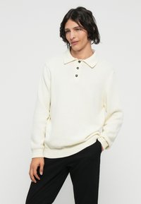 Weißen gestrickten Pullover mit Polokragen, der drei dunkle Knöpfe und gerippte Bündchen aufweist. Kombiniert mit schwarzen Hose.