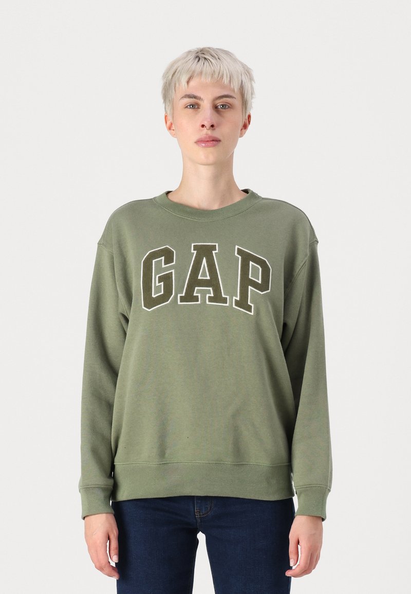Felpa verde oliva con testo bianco "GAP", girocollo, vestibilità rilassata, polsini e orlo a coste. Indossata con jeans blu scuro.