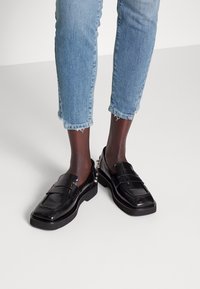 Czarne lakierowane loafersy na grubym spodzie z dekoracyjnym detalem, noszone z przyciętymi, jasnoniebieskimi dżinsami.