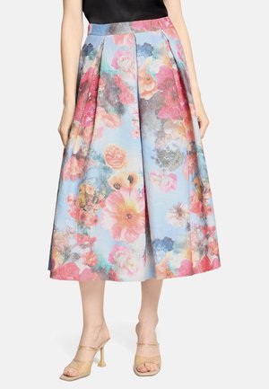 Kobieta w plisowanej spódnicy do połowy łydki z kolorowym kwiatowym wzorem i beżowych sandałach na wysokim obcasie.