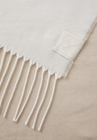 Vævet lysebrun tandet med hvide frynser. Har et lille, tekstureret mærke med et geometrisk design og initialerne "CK." Blød tekstur.