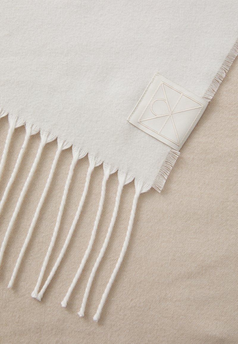 Woven light beige blanket with white fringes. Features a small, textured label with a geometric design and the initials “CK.” Soft texture.