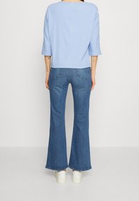 Donna che indossa un top azzurro chiaro, jeans flare a vita alta blu e sneakers bianche, in piedi con la schiena rivolta verso la macchina fotografica su uno sfondo bianco.