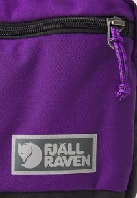 Sac à dos en tissu violet avec des accents noirs, doté d'une fermeture éclair et d'un badge logo en argent de Fjällräven à l'avant. Texture lisse.