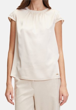 Frau trägt eine kurzärmelige, hellbeige Satinbluse und passende Hose, steht mit entspannten Armen an den Seiten.