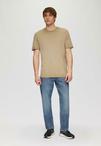 T-shirt beige à manches courtes avec un col rond, associé à un jean en denim bleu clair. Le modèle porte des baskets noires et se tient devant un fond uni.
