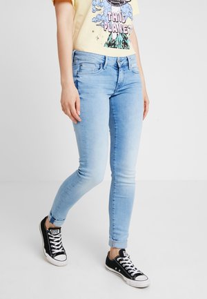 Persona que lleva puestos jeans ajustados azul claro, zapatillas negras bajas y una camiseta amarilla con estampado y el texto "Este Planeta".