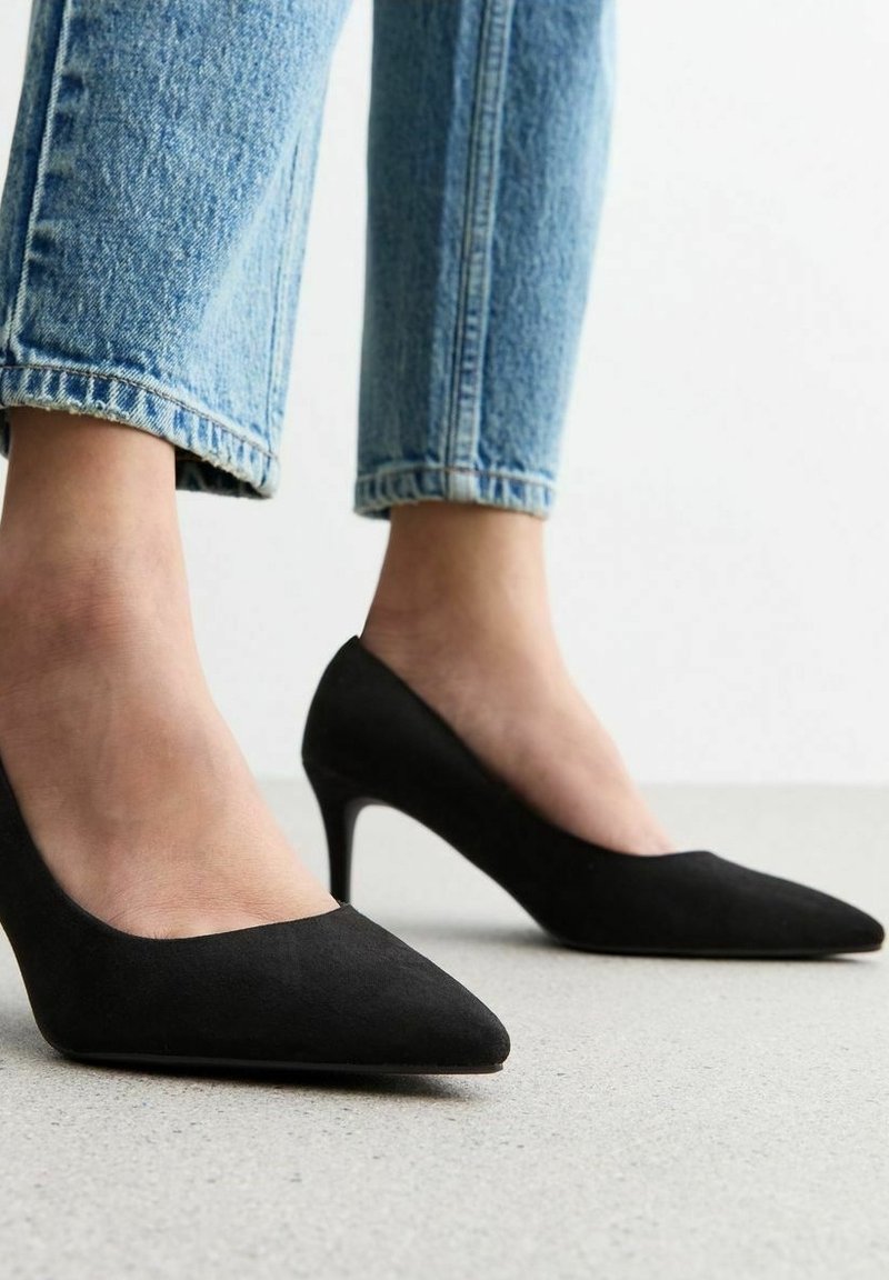 Pieds portant des escarpins noirs à bout pointu, associés à un jean bleu clair retroussé, posés sur un sol clair avec un fond blanc.