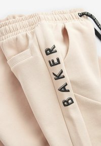 Beige sweatpants med elastisk talje, sidelomme og sort trykt tekst, der står "BAKER" langs venstre ben i fed skrift.