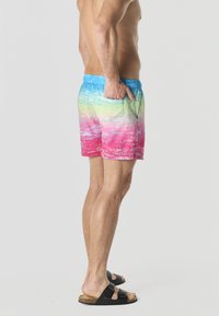 Homme torse nu en short de bain à dégradé coloré, main dans la poche, portant des sandales noires à boucle, debout sur un fond clair.