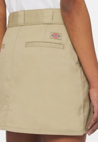 Gonna in denim khaki con vita aderente, tasche posteriori visibili e patch del logo Dickies. Taglio dritto con una texture liscia.