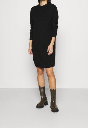 Frau in einem schwarzen, langärmligen Strickkleid und dunklen, groben Stiefeln bis zur Mitte der Waden steht vor einem einfachen hellen Hintergrund.