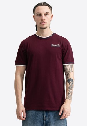 Uomo con capelli intrecciati che indossa una maglietta Lonsdale color mogano e jeans blu, mostrando il braccio destro tatuato e un'espressione neutra.
