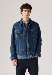 Donkerblauwe denimjack met drukknoopsluiting, twee borstzakken en studaccenten op de schouders. Gelaagd over een lichtere denimshirt.