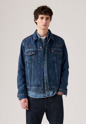 Veste en jean bleu foncé avec des boutons-pression, deux poches poitrine et des accents cloutés sur les épaules. Superposée à une chemise en jean plus claire.