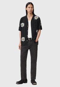 Czarna koszula z krótkim rękawem z białym kwiatowym haftem na ramionach, połączona z ciemnymi jeansami i czarnymi butami, o prostym wzornictwie.
