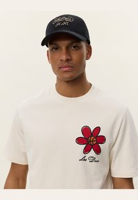 Camiseta de algodón blanca con un diseño de flor roja en el centro y un balón de baloncesto, con "Leo Deus" bordado en negro. Gorra negra con texto en dorado.