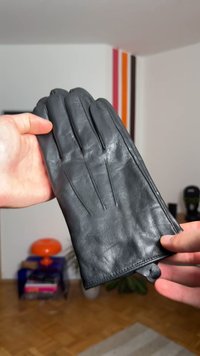 Deux mains tenant un gant en cuir noir avec des détails cousus dans une pièce avec un sol en bois et une décoration murale rayée colorée.