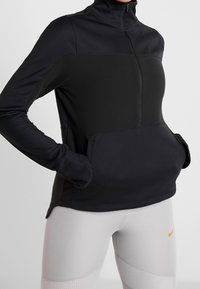 Sort atletisk pullover med høj krave, lynlås foran og sidelommer. Fremstillet af glat, elastisk materiale med en kontrasterende øverste panel.