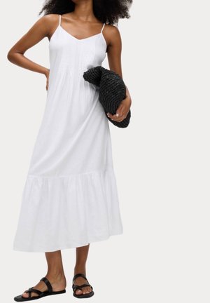 Kvinde iført en hvid ærmeløs midikjole, der holder en sort strikket clutch og bærer sorte flade sandaler.
