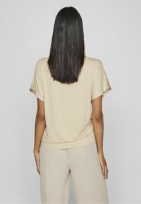 Beige kortärmad topp i mjukt tyg, med avslappnad passform och rund halsringning, visad bakifrån mot en neutral bakgrund.