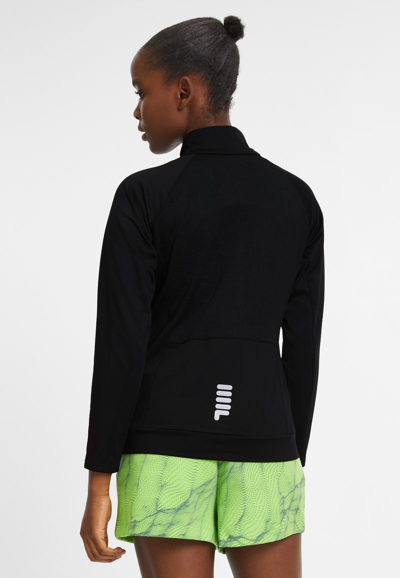 Donna con i capelli raccolti in uno chignon, indossa una giacca nera a maniche lunghe con un logo bianco sulla parte bassa della schiena e pantaloni corti a fantasia verde brillante.