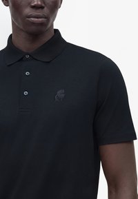Homme portant un polo noir à manches courtes avec trois boutons et un petit logo brodé sur la poitrine.