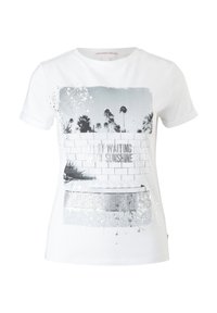 T-shirt blanc à manches courtes avec une impression en niveaux de gris représentant des palmiers, un mur en briques, et le texte « Il attend dans le soleil. »