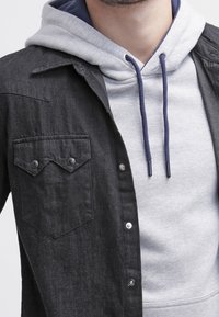 Mężczyzna ubrany w jasnoszary hoodie z niebieskimi sznurkami pod czarną, zapinaną na guziki kurtką z kieszenią na piersi i metalowymi napami.