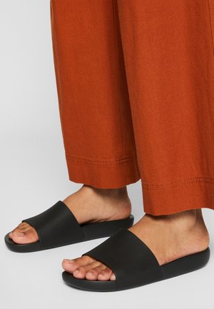 Personne portant un pantalon ample couleur rouille et des sandales noires à enfiler, debout sur un sol gris clair. Seules les jambes inférieures et les pieds sont visibles.