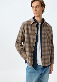 Rutet flannel skjorte i beige og brune nuancer med en brystlomme, knaplukning og lagdelt over en marineblå skjorte. Denim jeans.