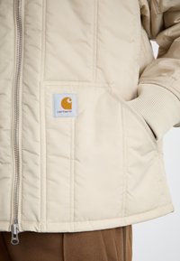 Beige gesteppte Jacke mit einem Reißverschluss vorne, gerippten Bündchen und einer aufgesetzten Tasche mit einem blauen Etikett und einem orangefarbenen Logo auf der Brust.