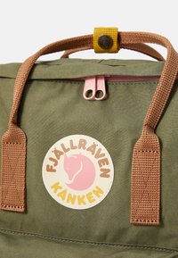 Zöld Fjällräven Kånken hátizsák barna fogantyúkkal, rózsaszín cipzárhúzókkal és kerek logóval, amelyen róka design és a márkaneve szerepel.