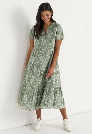 Kvinde smiler, iført en grøn blomsterprint kortærmet midi kjole og hvide sneakers, står op ad en hvid væg.
