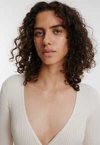 Rippstrick Pullover in Cremé mit tiefem V-Ausschnitt. Hat lange Ärmel und eine weiche Textur, die das figurnahe Design betont.