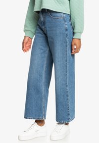 Jeans in denim blu a gamba larga con vita alta, caratterizzati da un design pulito e da dettagli minimi, abbinati a sneakers bianche.