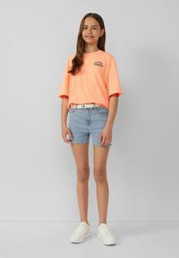 Pomarańczowa neonowa, oversize'owa koszulka z czarn�ą grafiką, zestawiona z jasnoniebieskimi szortami dżinsowymi i białymi trampkami. Prosty, casualowy design stroju.