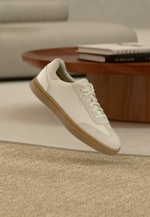 Λευκό αθλητικό παπούτσι χαμηλό με ταμπά σόλα, τοποθετημένο σε ένα μπεζ δωμάτιο με ξύλινο τραπέζι και στοίβες βιβλίων στο φόντο.