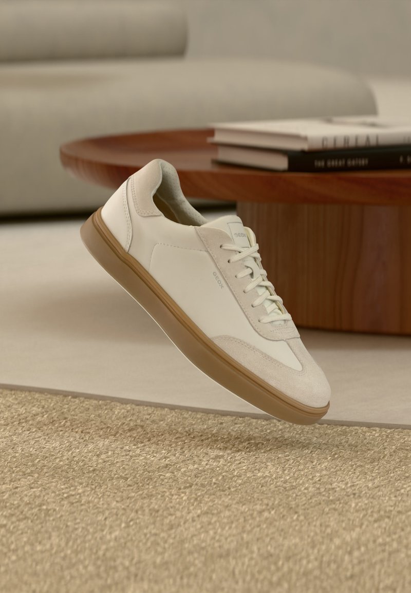 Λευκό αθλητικό παπούτσι χαμηλό με ταμπά σόλα, τοποθετημένο σε ένα μπεζ δωμάτιο με ξύλινο τραπέζι και στοίβες βιβλίων στο φόντο.