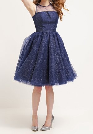 Vestido azul marino con un torso ajustado y una falda de tul acampanada. Presenta detalles brillantes y un escote transparente. Combinado con tacones plateados.