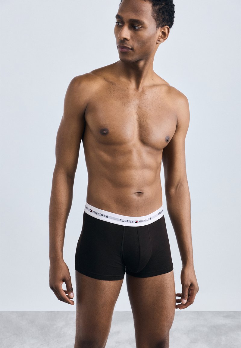 Boxer in cotone nero con elastico bianco a marchio, design aderente e texture liscia. Il modello posa in posizione eretta su uno sfondo neutro.