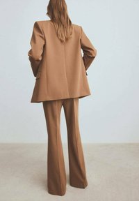 Bruine op maat gemaakte blazer met gestructureerde schouders, gecombineerd met bijpassende uitlopende broeken. Gemaakt van een soepele stof, met zijzakken.