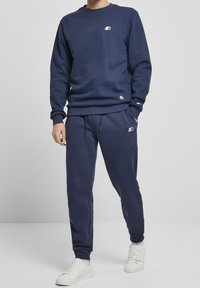 Hombre vistiendo sudadera azul marino y pantalones deportivos con zapatillas blancas, de pie contra un fondo gris claro liso.