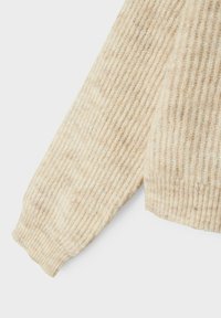 Pull en tricot beige avec une texture côtelée, des manches longues et une coupe décontractée, présentant des filaments plus foncés visibles à travers le tissu.