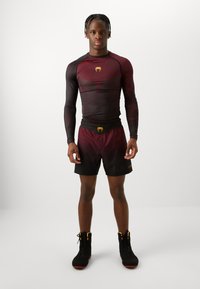 Μαύρη και μπορντό αθλητική στολή με μακρυμάνικο, εφαρμοστό τοπ με λογότυπο και σορτσάκι. Η υφή είναι λεία; τα παπούτσια είναι μαύρα με ψηλή γάμπα και κόκκινες λεπτομέρειες.