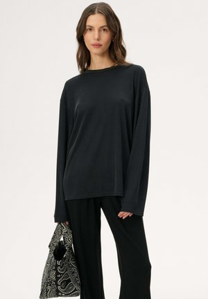 Kobieta ubraná w luźną czarną bluzkę z długim rękawem i spodnie, trzymająca czarną i białą torbę w wzory na jednolitym tle.