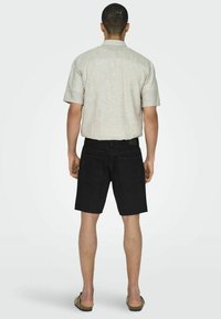 Camisa de lino de manga corta de color claro con cuello trasero, combinada con pantalones cortos de mezclilla negra y sandalias, vista desde la parte posterior.