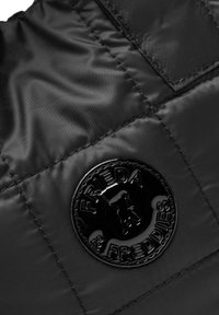Zwart gewatteerd puffer stof met een glanzend rond logo patch met de tekst "Frieda & Freddies". Gladde textuur met zichtbare stikdetails.